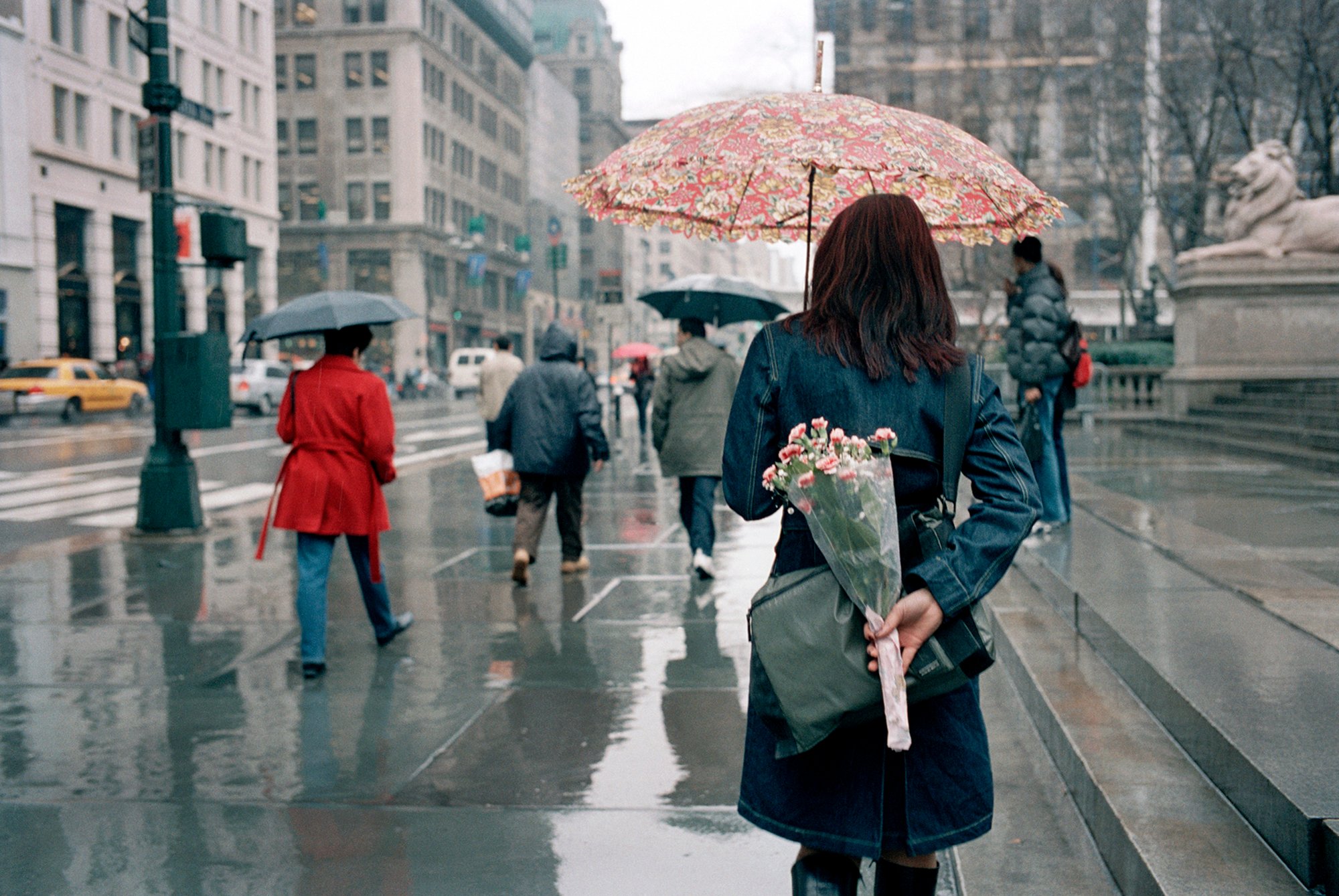 A person walking on a rainy street in front of the New York Public library, holding a flower printed umbrella and a bouquet of carnations behind their back.