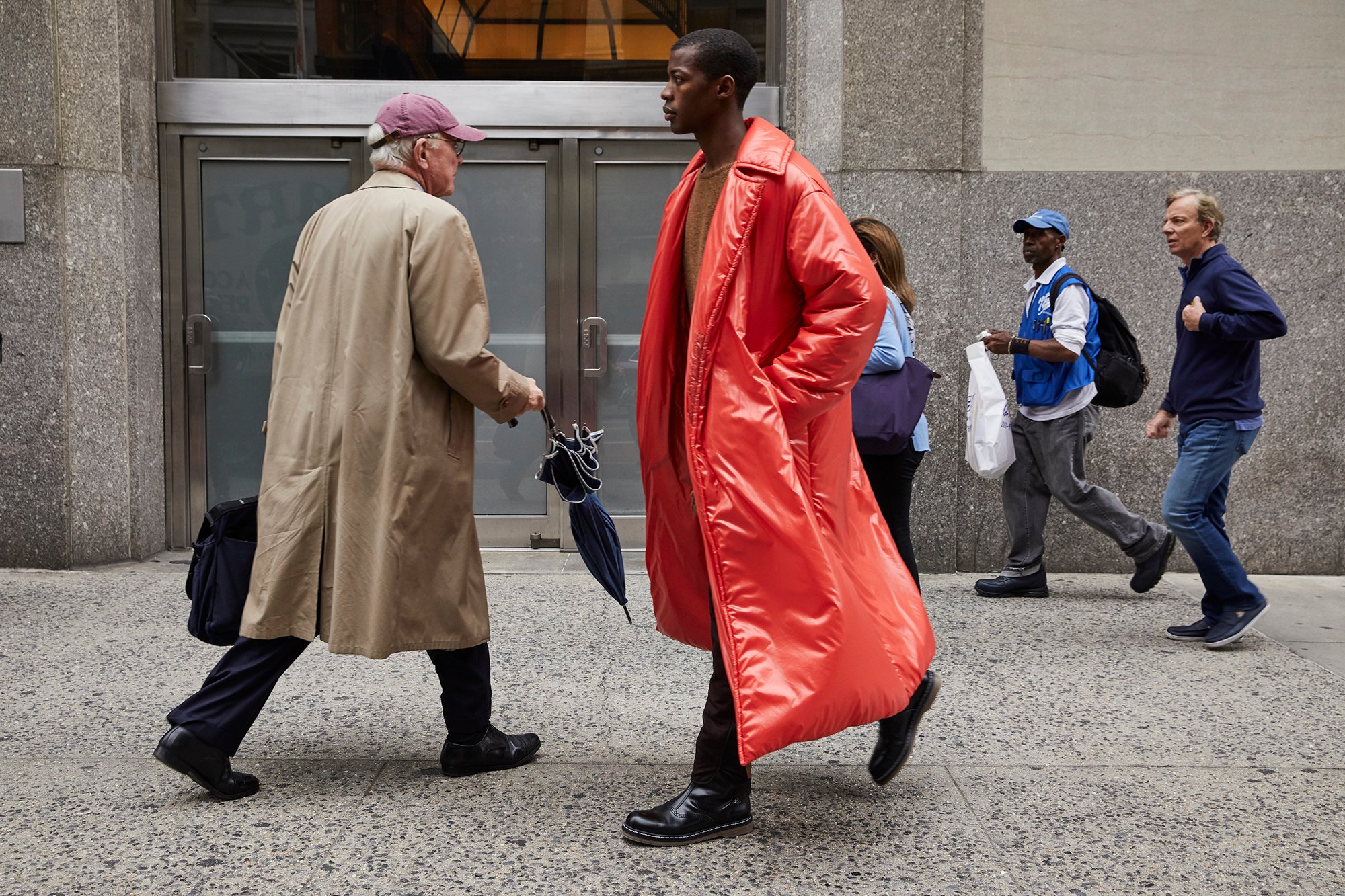 A young man walking with a shiny, smooth, puffy, plasticky scarlet trenchcoat. He walks forward while an older man in a typical beige cotton trenchcoat steps aside for him, shifting the weight of his closed umbrella away.