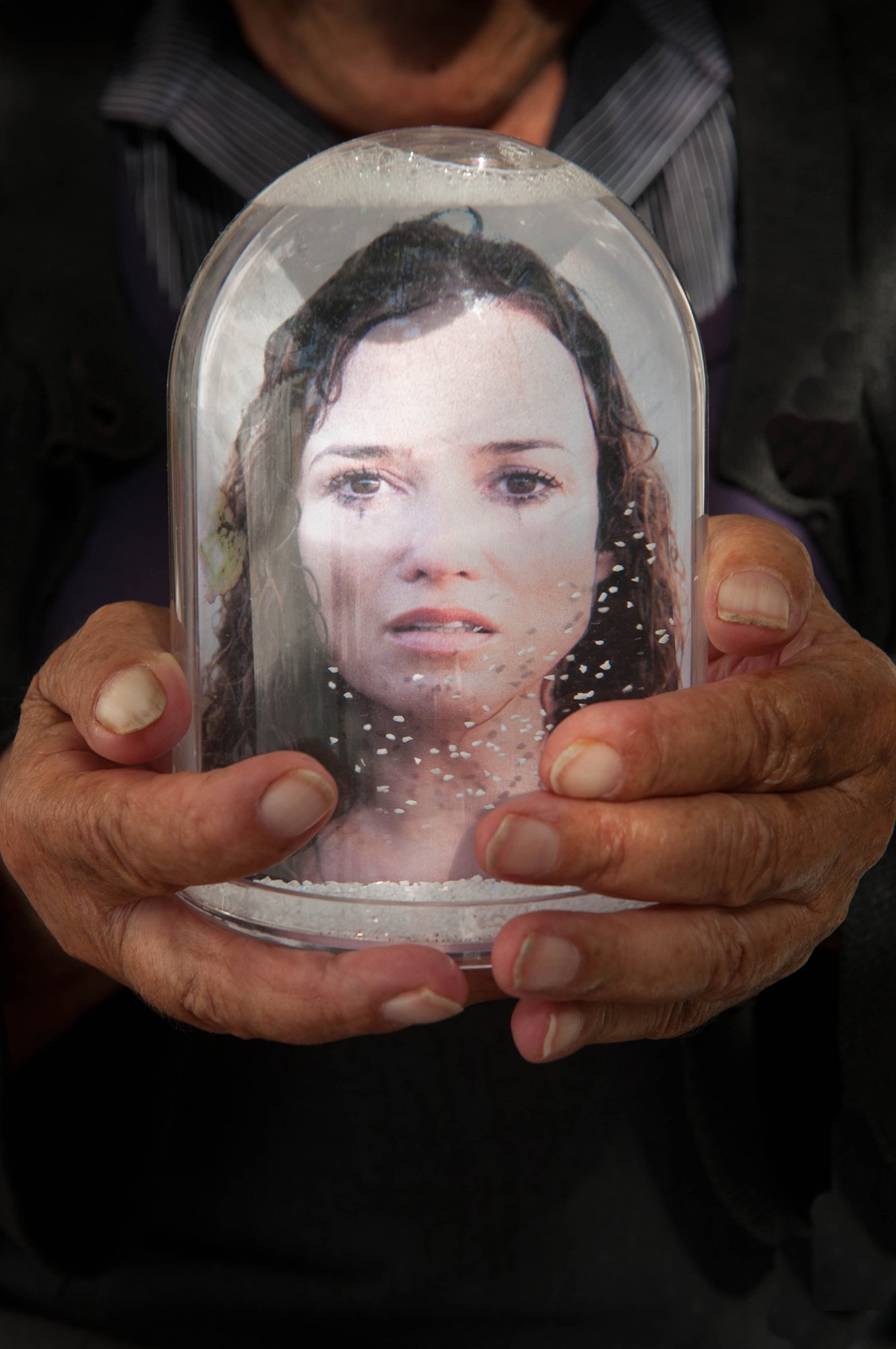Old hands with dirty thumbnails holding a snowglobe with a large print of a woman's distressed, possibly crying face.