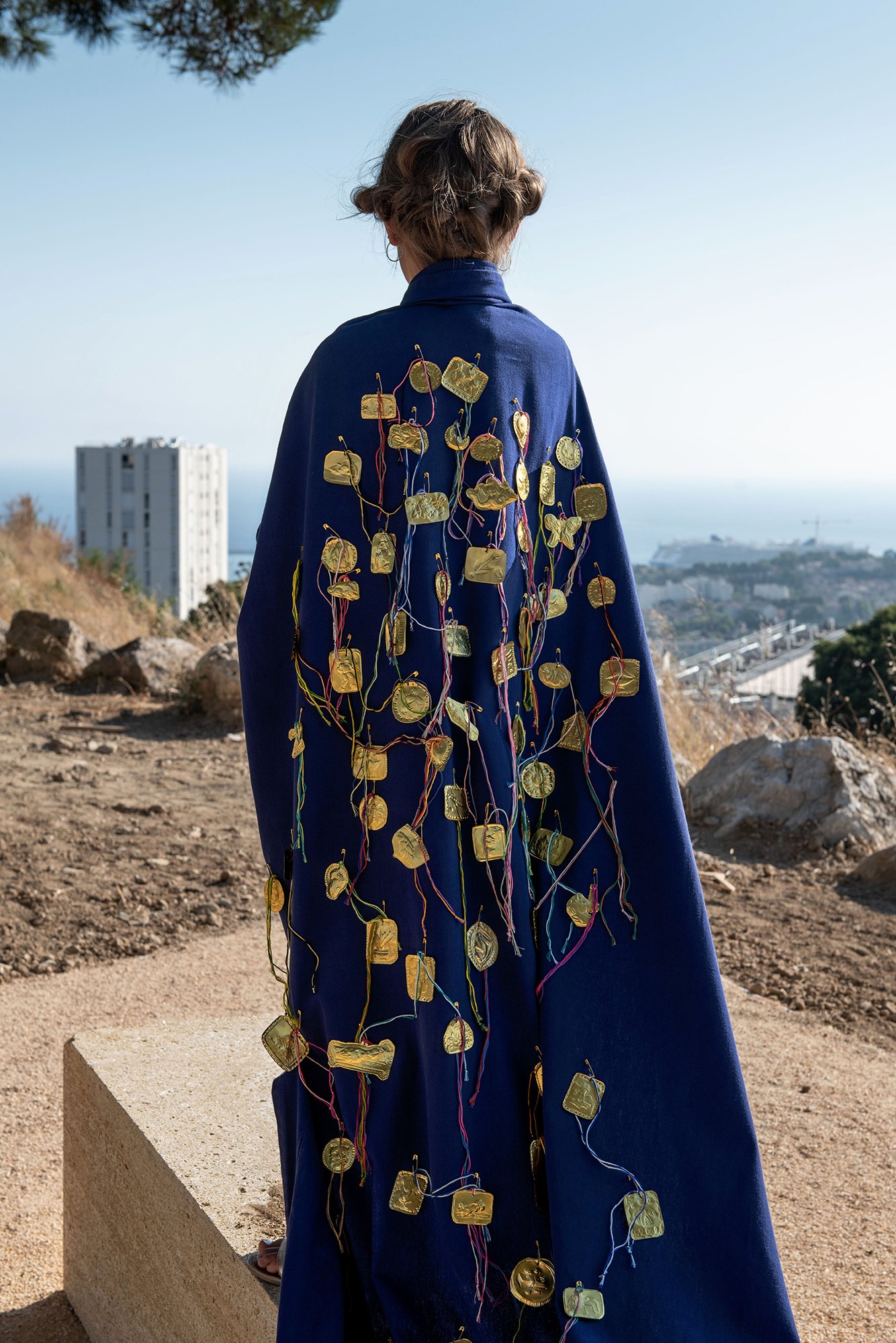 A person standing on a stone overlooking lower ground of a town by the sea. The person is pictured wearing a deep blue coat with dozens of gold medallions pinned to their back, and multi-colored threads streaming out from underneath.