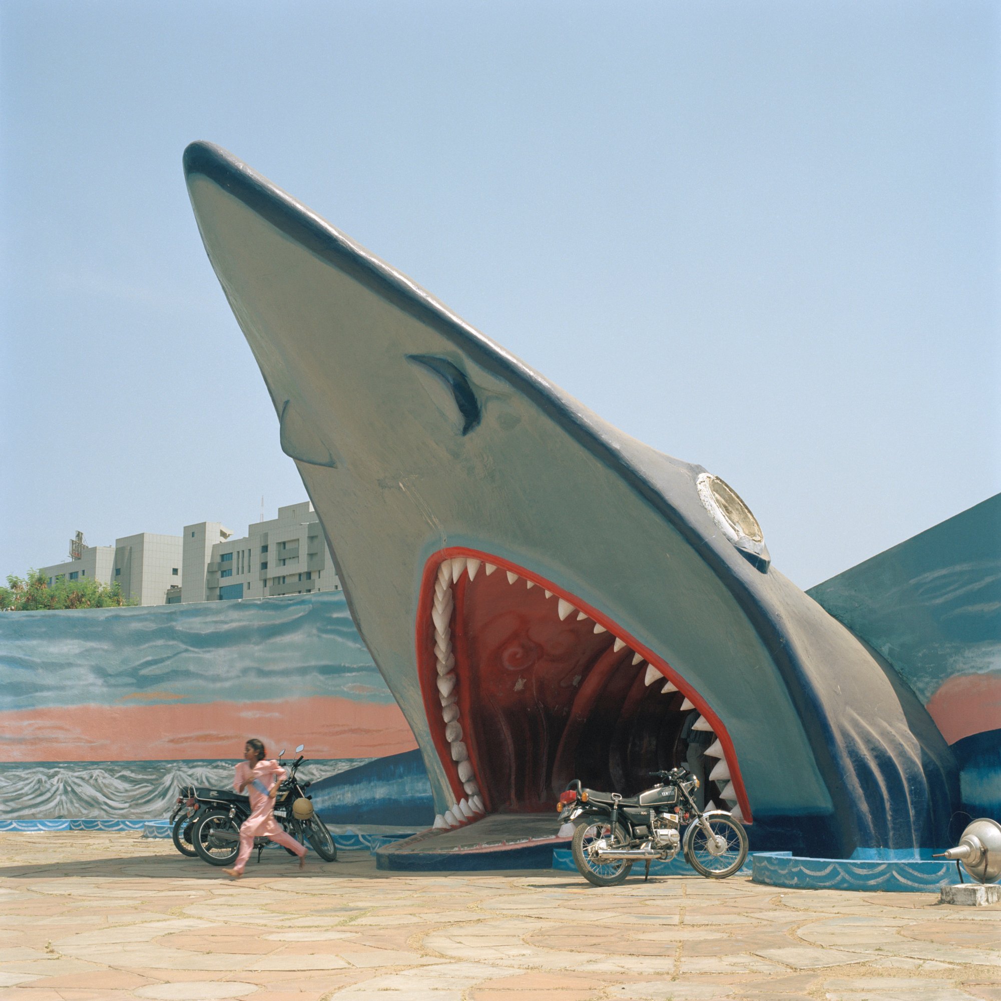 A person running between parked motorcycles, in the shadow of a giant sculpture of a shark's head emerging from the ground with its mouth open as a portal.