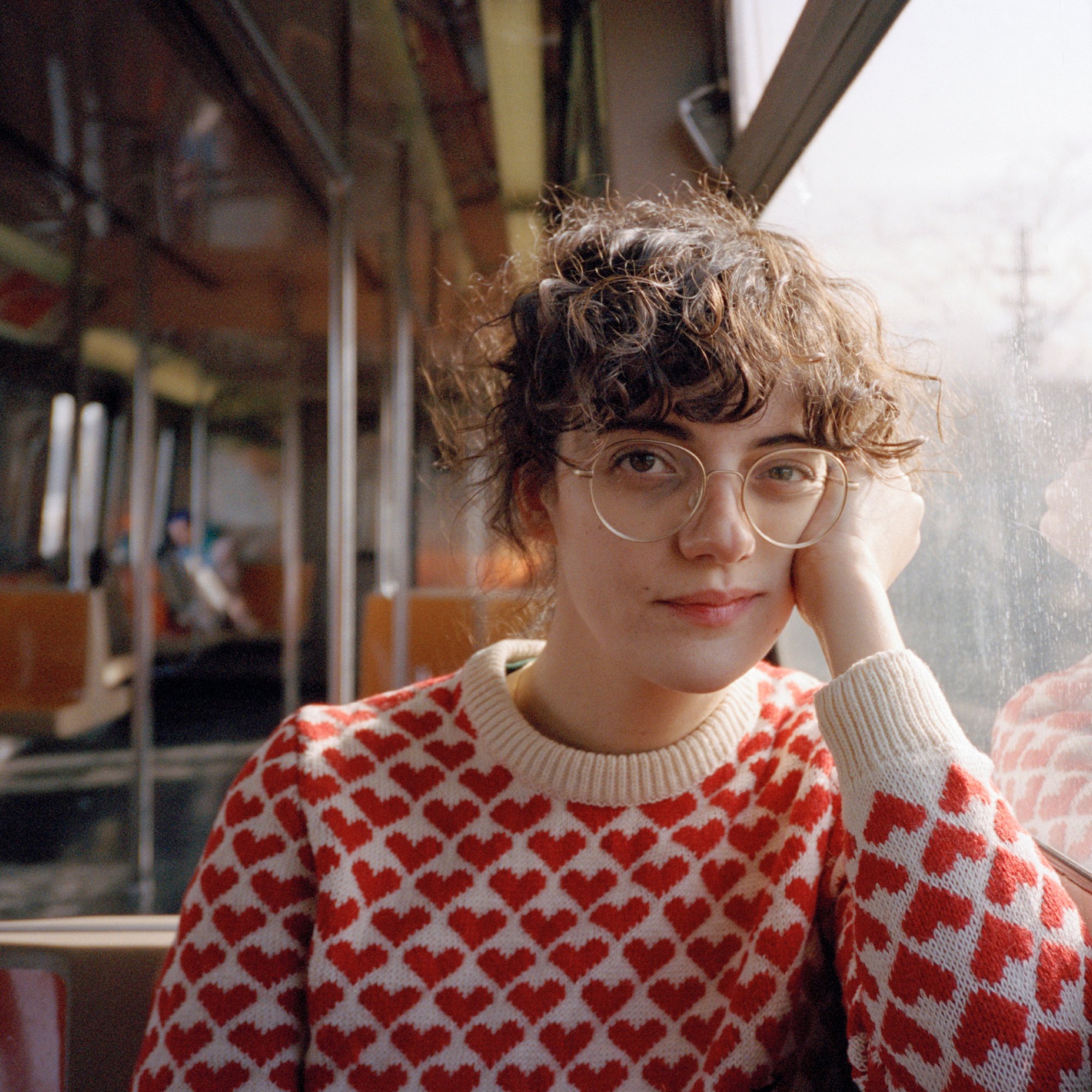 A portrait of a person with light skin, dark curly hair, glasses, sweater, sitting in a train car. Their left hand is holding their head up and pulling their cheek back into a very slight smile.