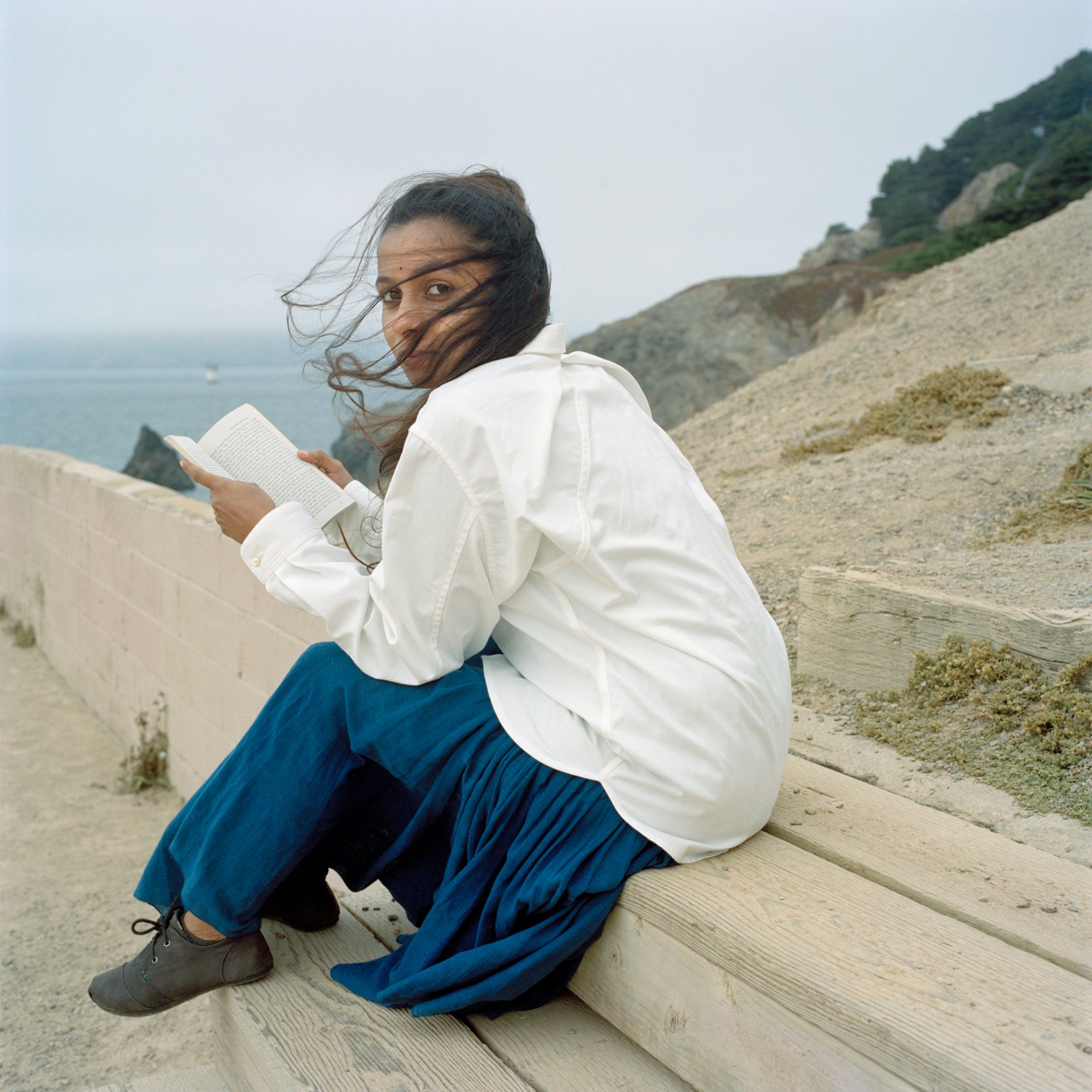 A person holding a book and sitting outside on sandy terrain overlooking an ocean.