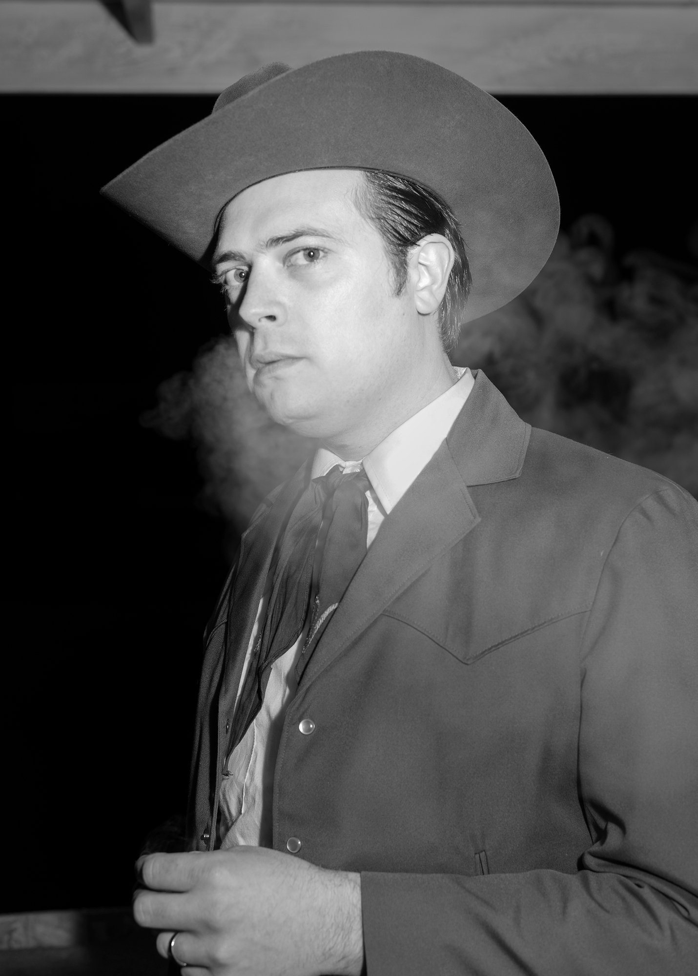 A portrait of a person with short slicked back hair underneath a suede hat with a large brim, surrounded by a little fog near the mouth.