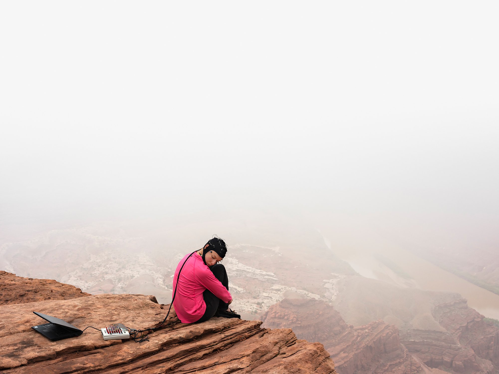 A person sitting and hugging their legs at the edge of a rocky cliff overlooking a foggy dry and mountainous landscape. The person is wearing what appears to be an EEG wired to a laptop.