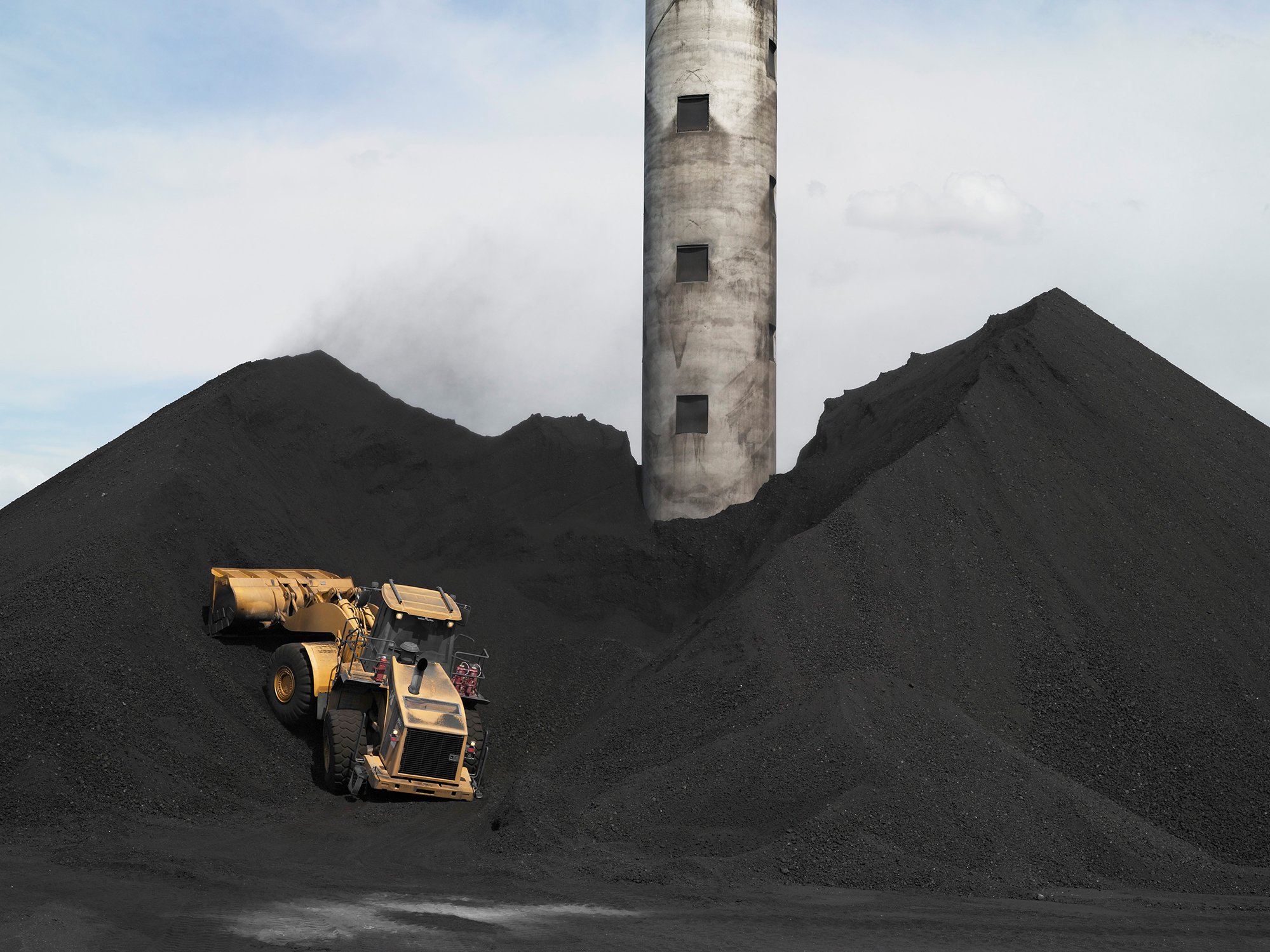 A scene of mountains of black soot. A concrete tower emerges from the pile and dirty front loader truck is climbing up the pile.
