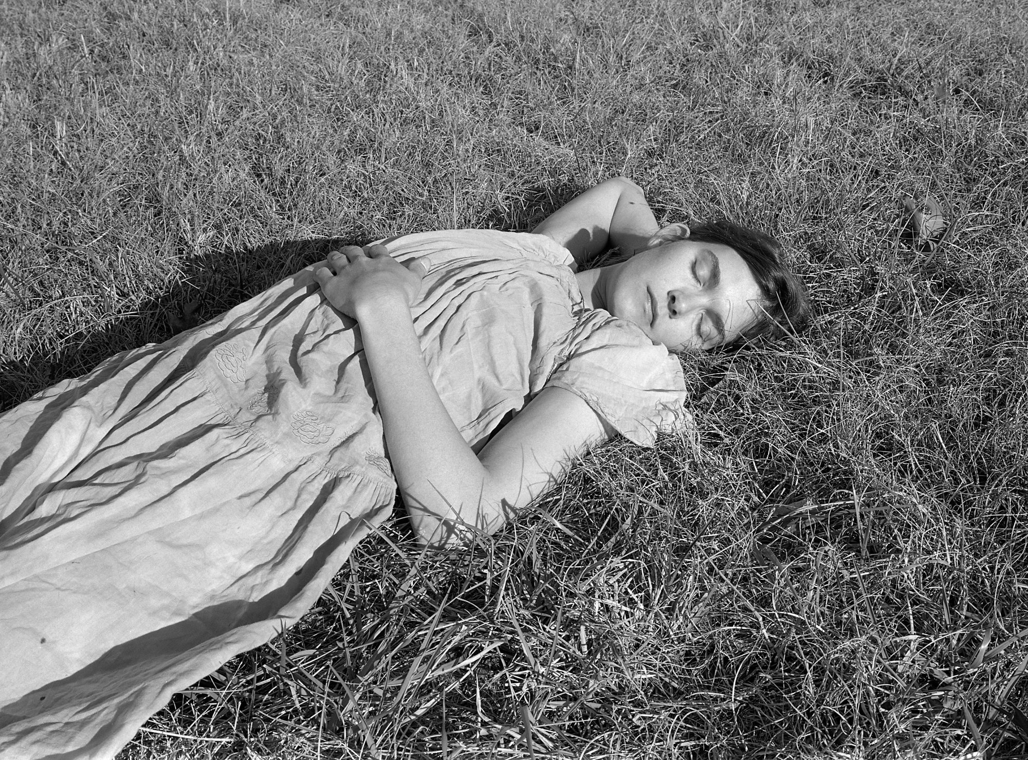 A woman with light skin, in a wrinkly linen dress lying in the grass, eyes closed, one hand over her belly and the other under her head.