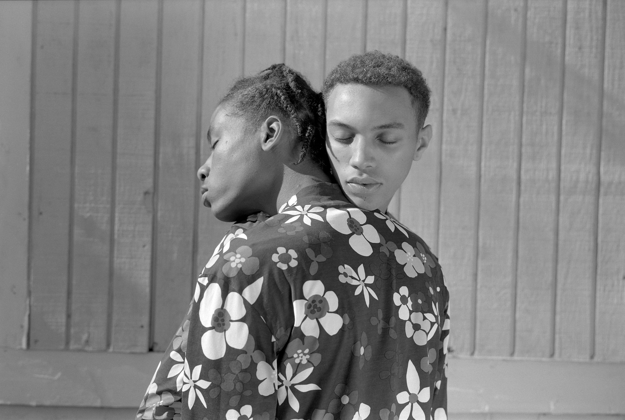 Two young people with dark skin in a sort of embrace, with eyes closed. Both are wearing shirts with abstracted floral illustrations. One has their back turned to the camera and head turned to the side, and the other is resting their head on the shoulders of the first one, facing the camera. 