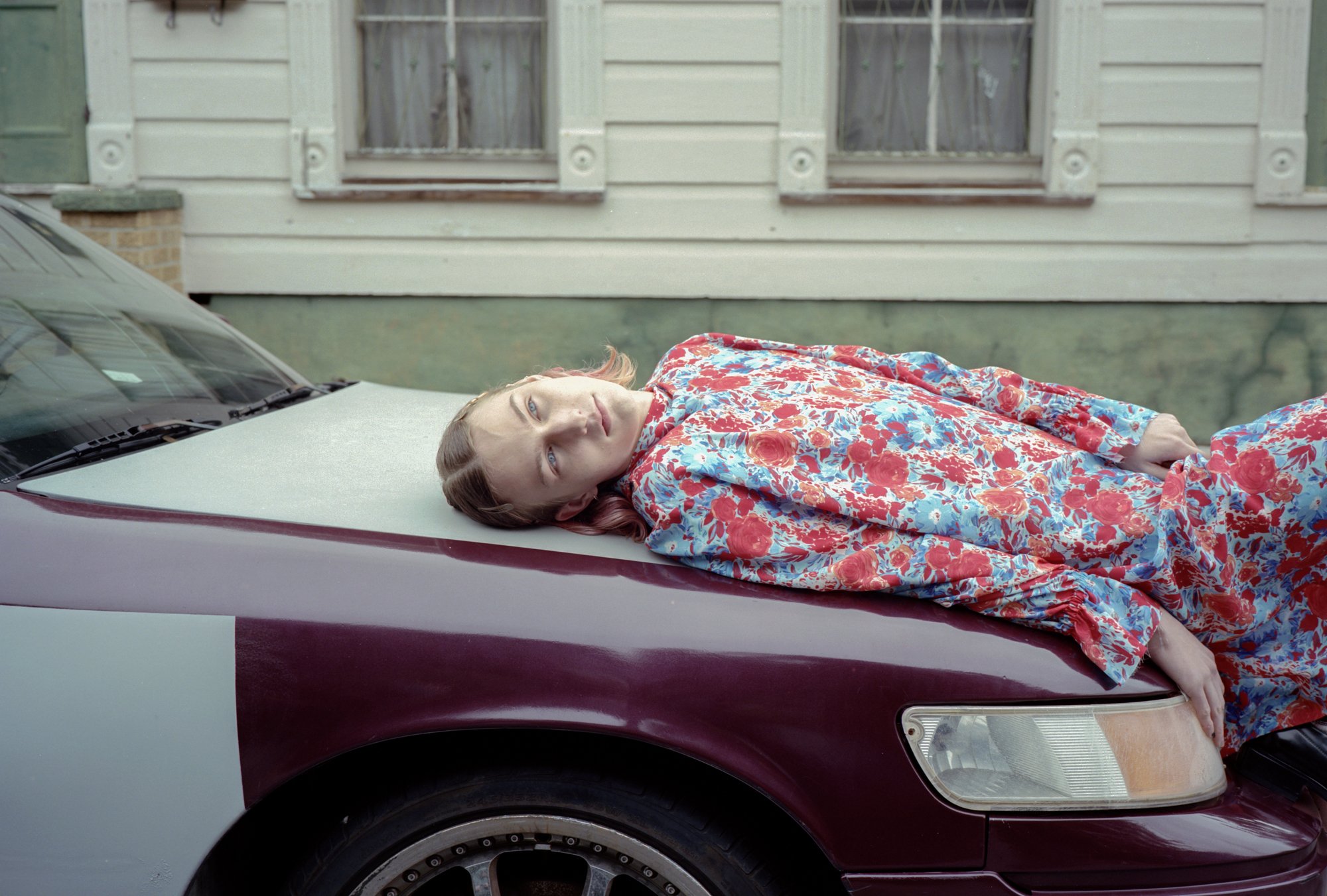 A person with light skin, long combed hair curling at the ends, wearing a blue and red floral print dress with long sleeves covering long arms and torso that is melting over the glossy hood of a burgundy car. Their hand is just resting over the headlight.