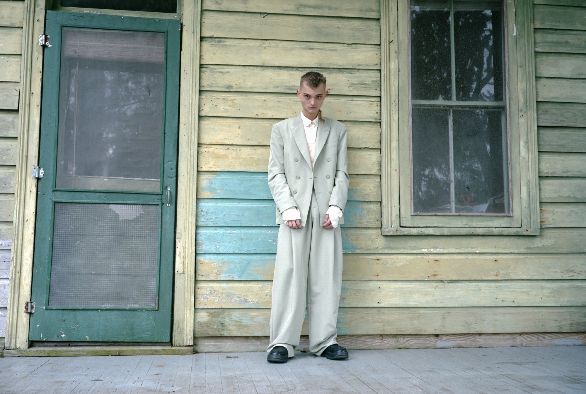 On the porch of a seemingly old house with worn wooden siding, stands a person with light skin, short brown hair, and a seemingly skinny frame, wearing an oversized light greyish beige suit.