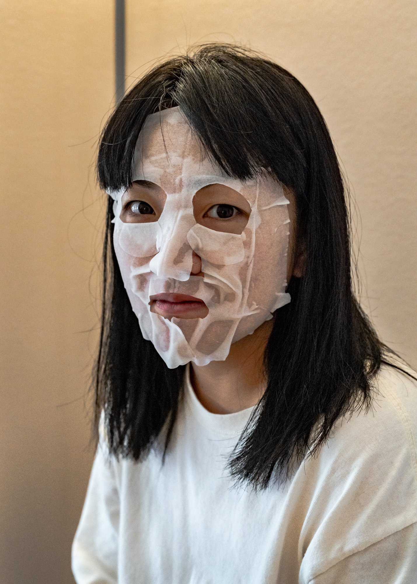 A person with long black hair wearing a disposable skin care face mask with cutouts for their eyes and mouth. Their banks fall in front of the face mask over their forehead, and they have a rather neutral face with a bit of a hunch in the shoulders. 