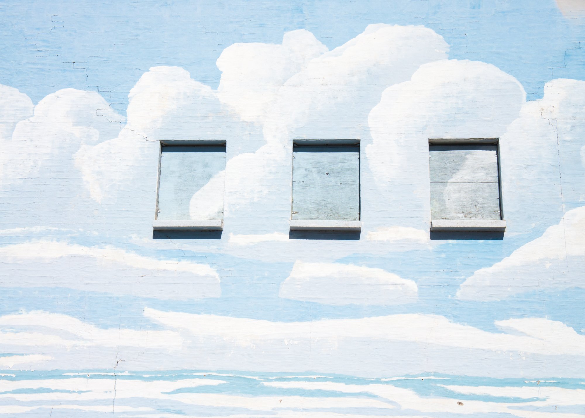 A brick wall painted like a blue sky with large white clouds. The wall has three windows boarded up with plywood that have also been painted over.